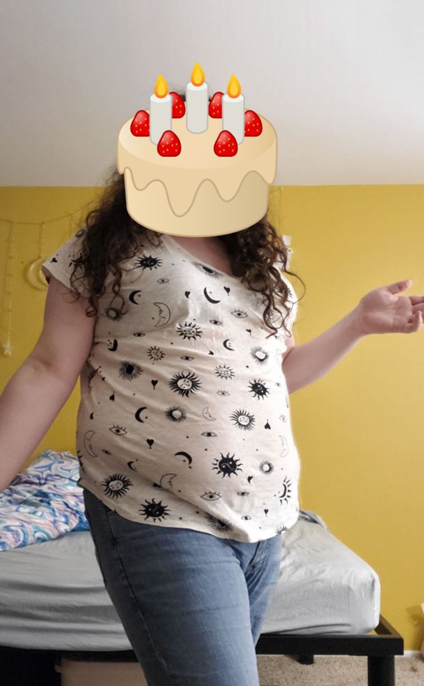 A medium wide shot of the same girl in a shirt decorated with suns and moons in front of a bed frame. She has a cake emoji covering her face