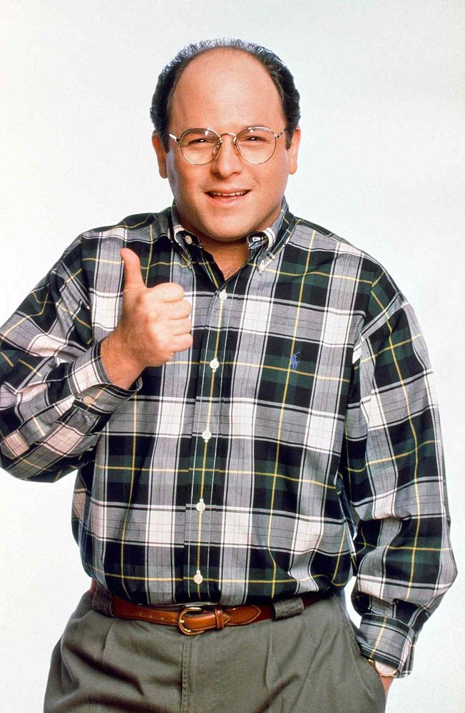 Actor Jason Alexander posing with a thumbs up. He's wearing a plaid shirt and tan pants with a brown belt and gold rimmed glasses. He stands in front of a white background.