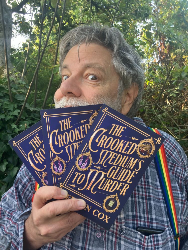 Outside, trees background. Stephen Cox vaguely manic looking with grey hair and beard with a fan of three paper copies of The Crooked Medium's Guide to murder. A rich dark blue background, and a touch of mist. The text appears in gold in a gold rectangular frame, the font is old fashioned implying C19th century. The frame has three rondels or cameos each containing a picture. Top right is a golden bee brooch with the body and one wing streaked with red blood. Middle left is foxgloves, spires of small bell flowers in pink, white and purple. Bottom right is a dark brown medicine bottle, with a pale label. The word LAUDANUM is just visible. There are pink highlights on some of the letters. Taken together it is bold, mysterious, and Victorian. 