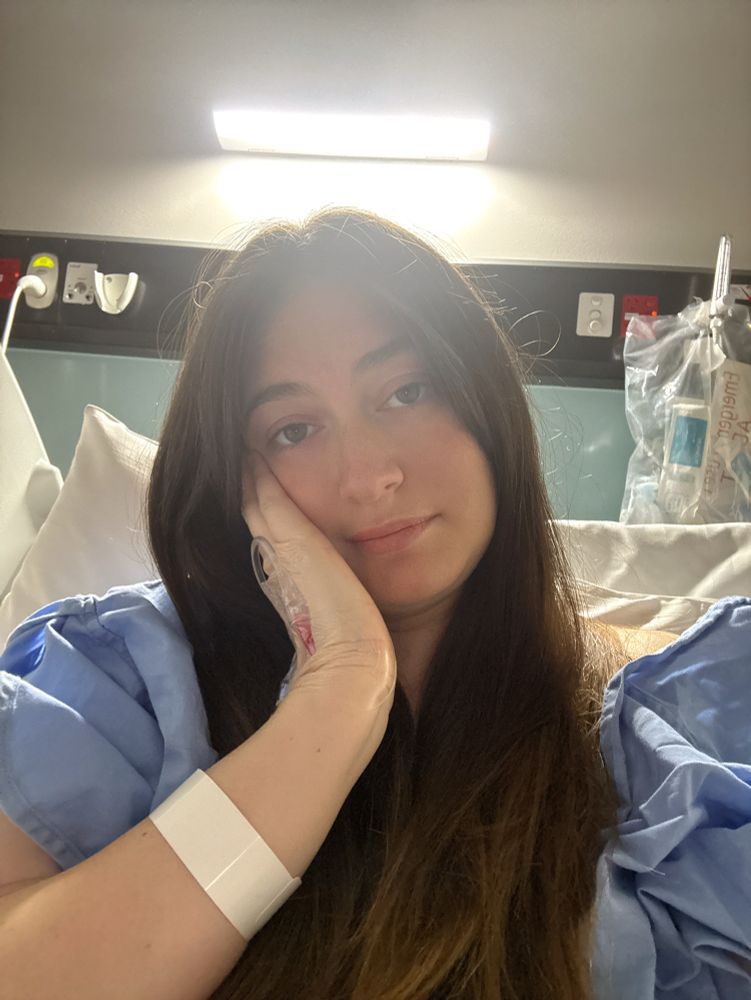 Brunette woman with long hair in a blue hospital gown in a hospital bed. Her right hand is resting on her cheek and she is looking at the camera with a tired but relieved expression.