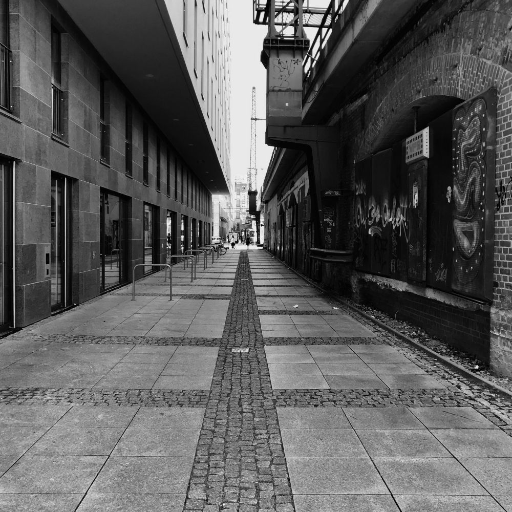 Schwarz-Weiß-Foto (Rohbild) eines Radwegs in Berlin parallel zur Hauptstraße auf dem Weg zum Savignyplatz. Links sind ein Hochhaus und metallene Anlehnbügel für Räder, rechts die Viaduktbögen der S-Bahn zu sehen. 