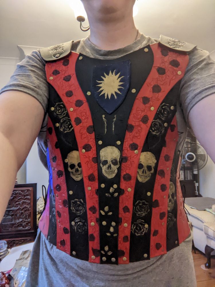 A torso shot of a person wearing black and red panelled leather armour over a grey t shirt. The armour is tooled with black roses on red panels, and gold skulls, roses and skeletons on the black panels. There is also a dark blue shield panel tooled with a golden sun at the top of the front panel.