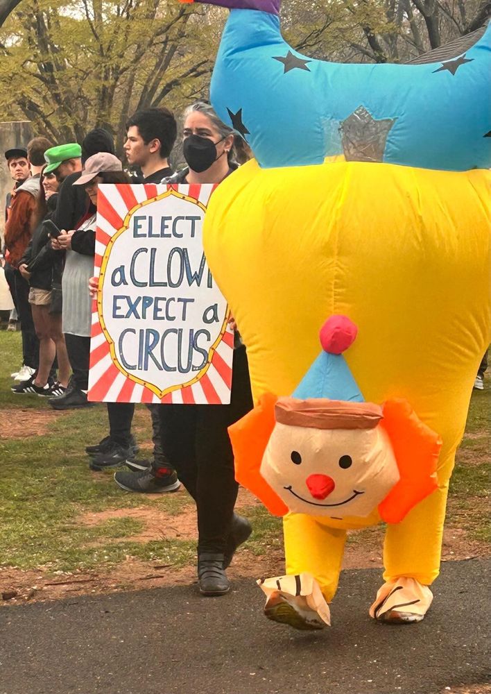 A protest sign says "Elect a clown, expect a circus" while a person in a blow up clown suit walks next to them