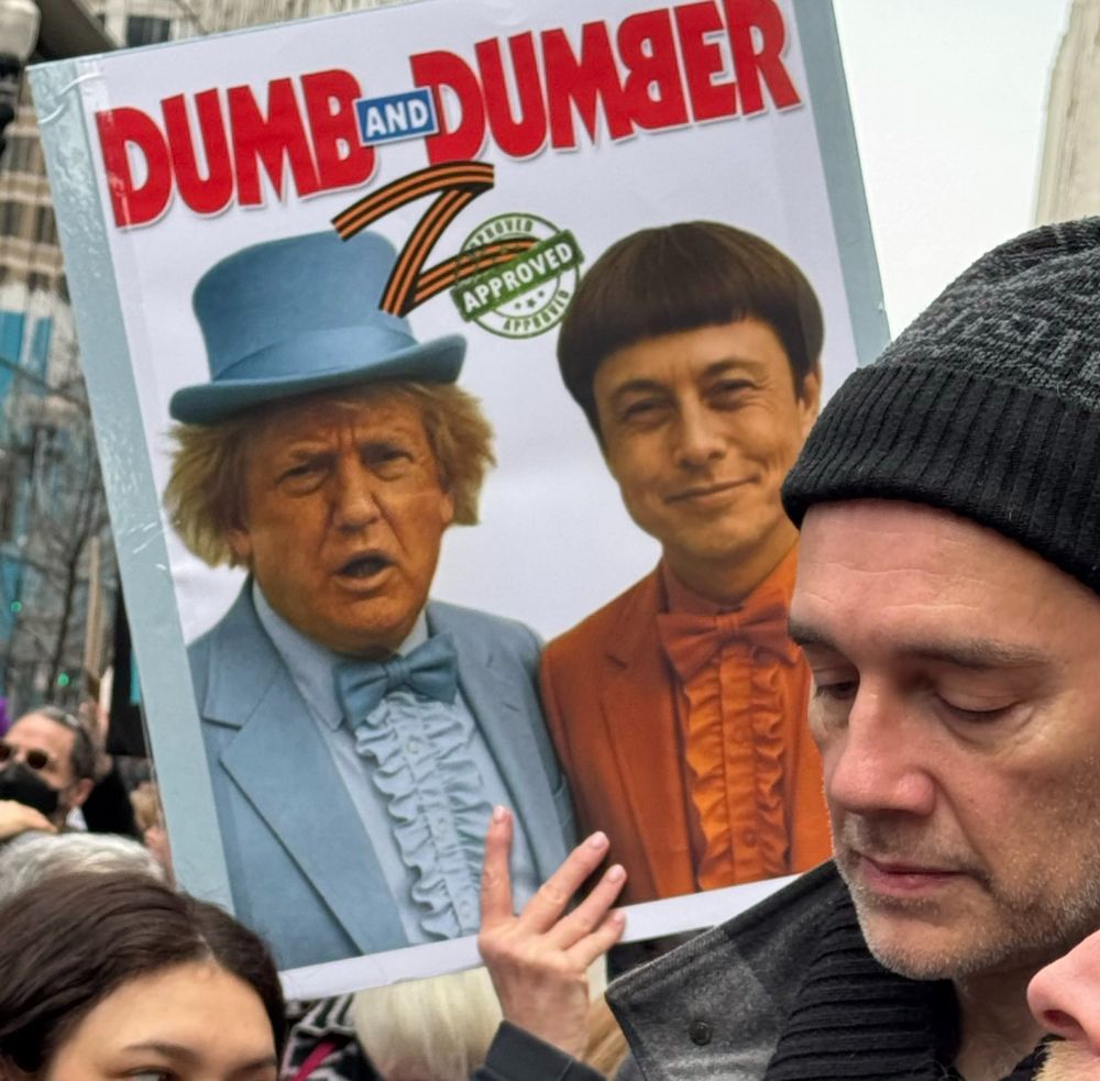 A protest sign parodying Dumb and Dumber the film, where Trump is in a blue suit like Jeff Daniels and Elon Musk is in orange like Jim Carrey