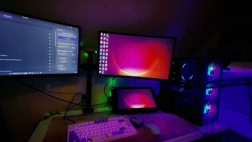 A PC setup. 2 monitors are on a mount clamped to the back of the desk, and a Huion Kamvas 13 is below the right monitor. The PC tower is on the right side of the desk, and a purple and white keyboard and blue mouse sit on a pink desk mat. A microphone arm is clamped to the far right side of the desk. 