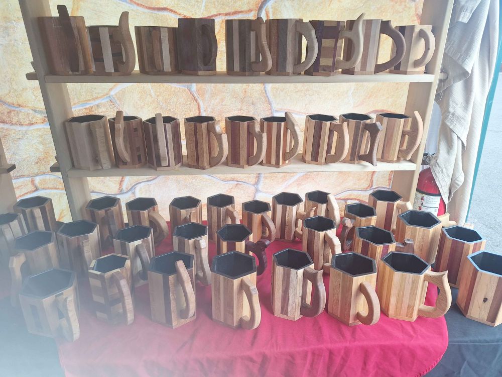 A table full of wooden mugs. Some sit on the black and red tablecloth, others are on a 2 tiered shelf to the back. They are made of various wood types and are all sealed on the inside with a black coating. 