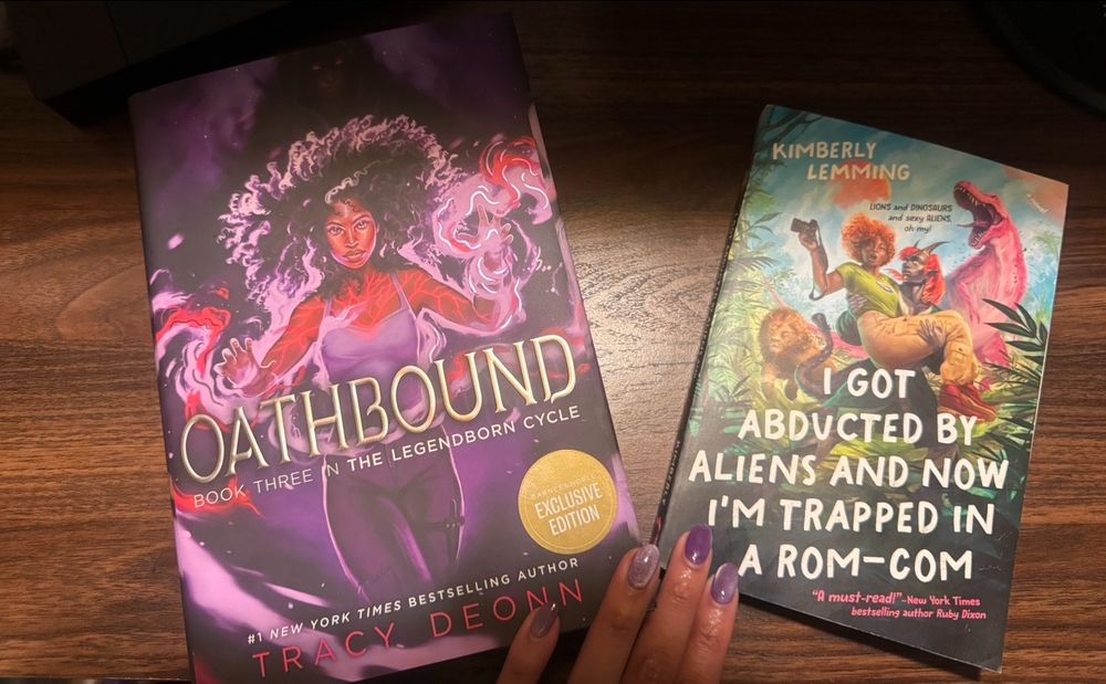 Two books on a desk, one purple (Oathbound by Tracy Deonn) one green (I Got Abducted by Aliens and Now I’m Trapped in a Rom-Com by Kimberly Lemming). The purple has a fierce looking black teen girl wielding purple and red magic. The green book has a red-headed black woman in the arms of a horned alien with a lion and a dinosaur in the background. 