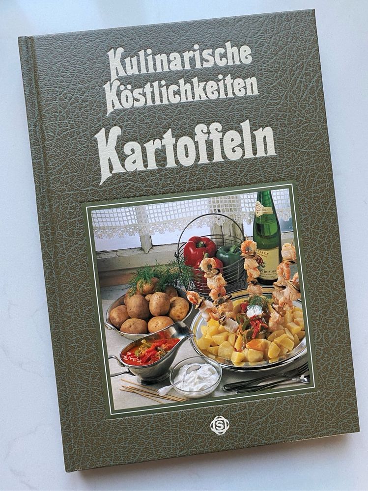 An olive green hardback German cookbook called Kulinarische Köstlichkeiten / Kartoffel. It has a very 1980s photograph of an arrangement of potato dishes on it. The book is lying on a pale grey kitchen counter.