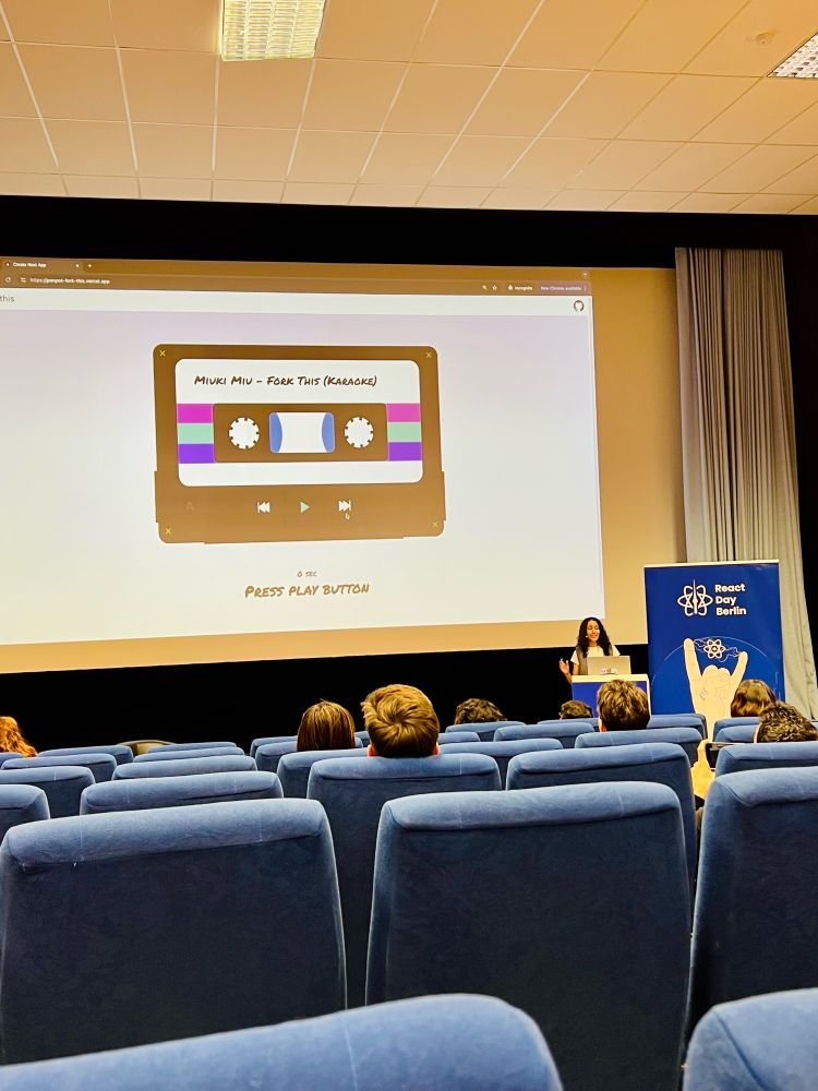 A presentation from Elizabet Oliveira at React Day Berlin. The speaker is standing at a podium and is presenting a slide showing a retro cassette tape design with the text “Miuki Miu - Fork This (Karaoke).” Below the cassette image, instructions say “Press Play Button.”