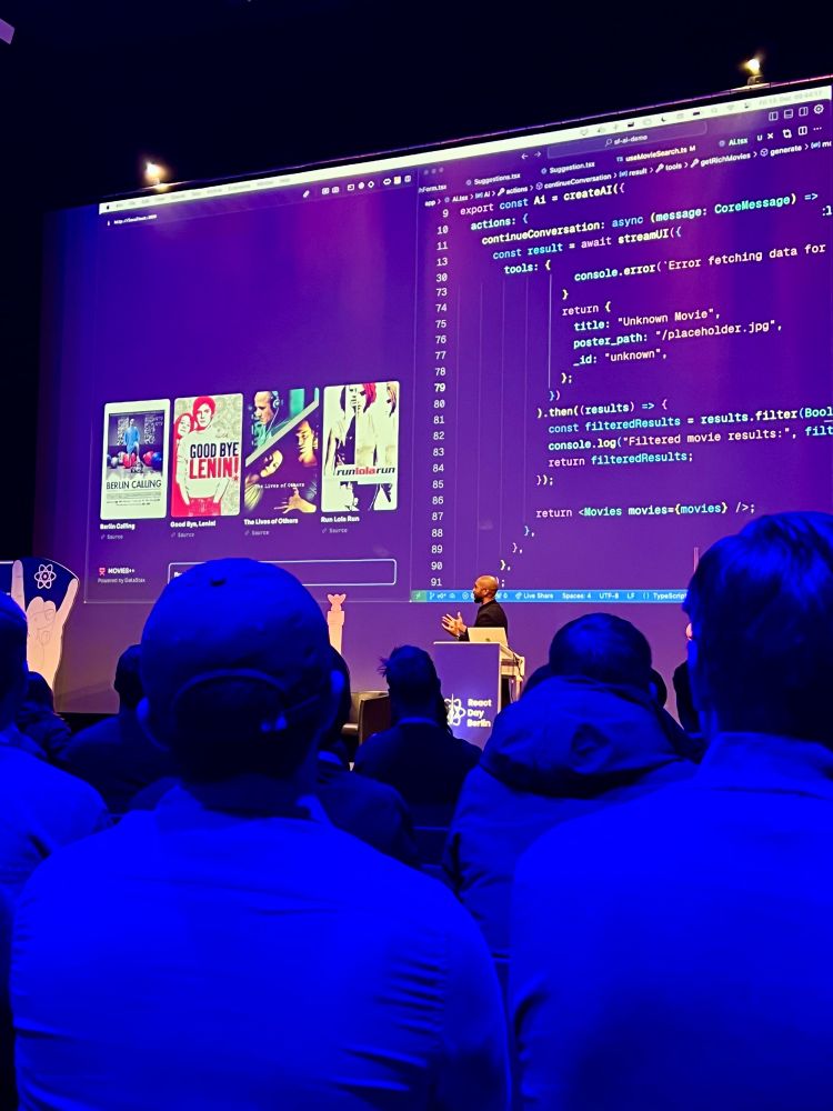 An auditorium filled with attendees watching Tejas Kumar present at React Day Berlin. The presenter is demonstrating a live coding session, showing a UI created by generative AI featuring movie posters like Berlin Calling, Good Bye Lenin!, The Lives of Others, and Run Lola Run. The room is illuminated with blue lighting, and the React Day Berlin logo is visible on the stage.