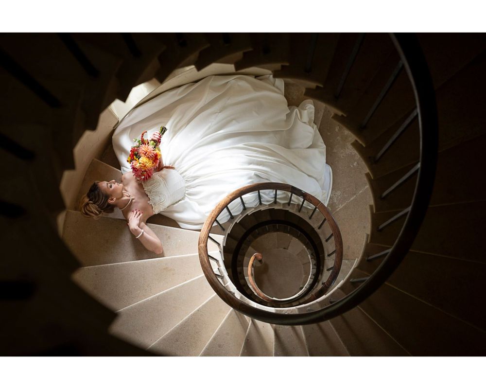 A woman in a long, white, satin wedding dress, holding a colourful bouquet, is see from above, lying backwards on a spiral stone staircase