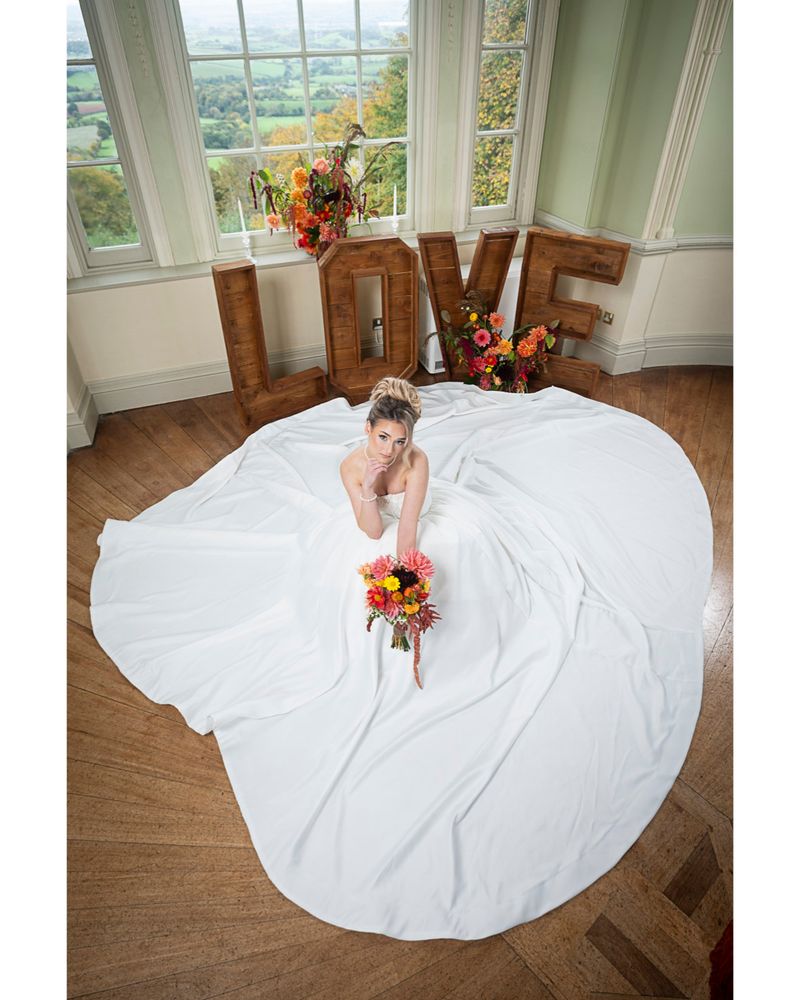 A woman in a long, white, satin wedding dress holding a colourful bouquet sits on the floor with the dress spread out around her. Behind her, wooden letters spell out the word 'love'