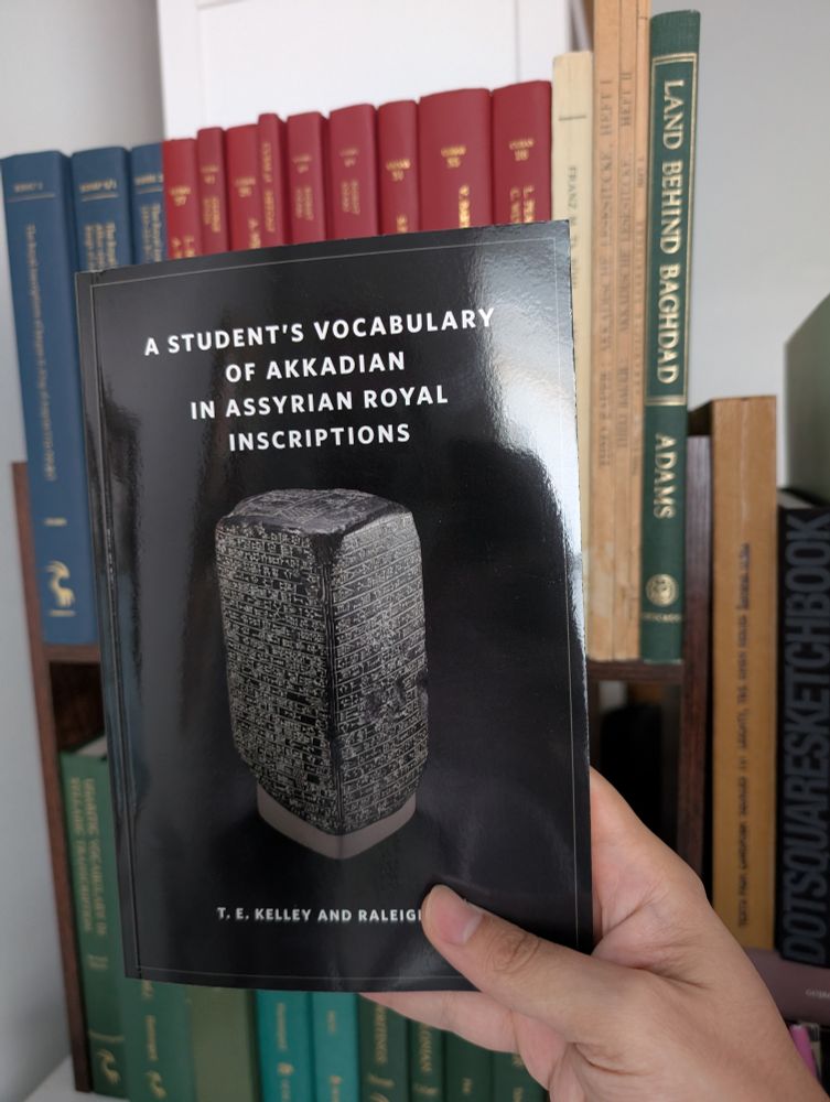 A hand holds up "A Student's Vocabulary of Akkadian in Assyrian Royal Inscriptions" by T.E. Kelley and Raleigh Heth.
