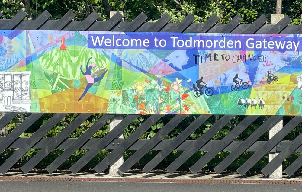 An advertising banner mounted on a fence at the back of a platform, which says “Welcome to Todmorden Gateway”.