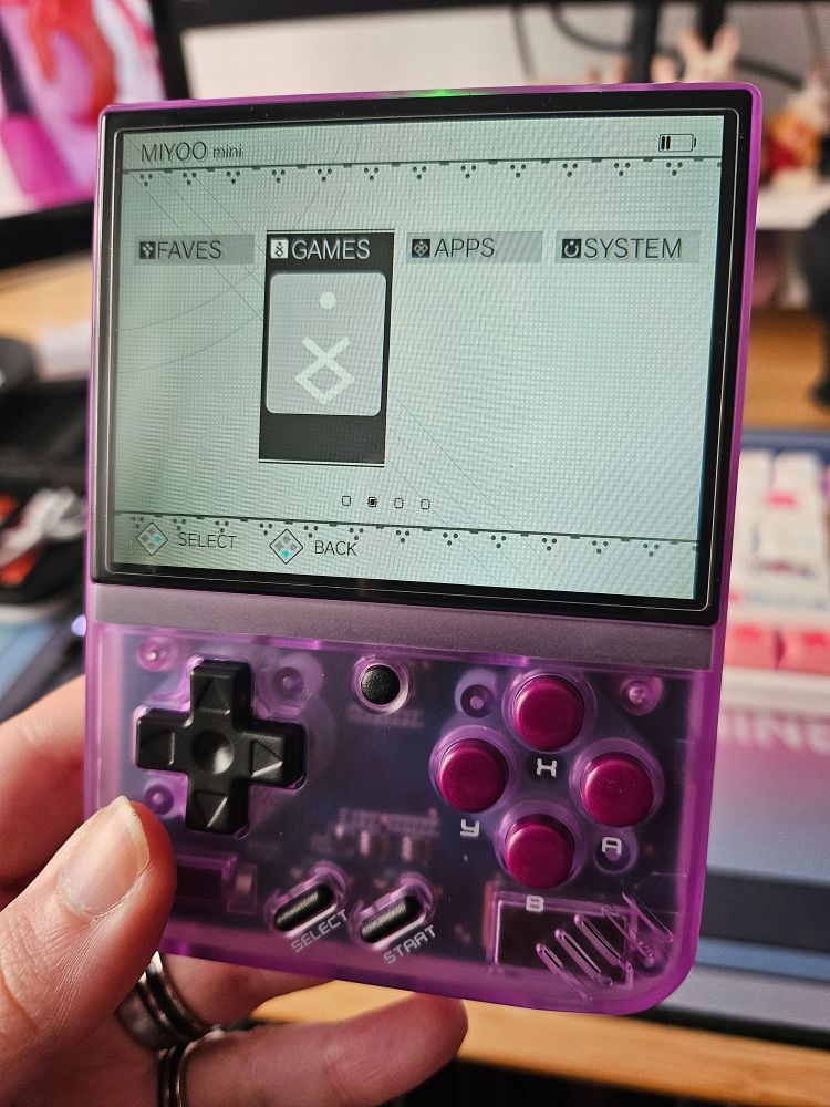 Photo où je tiens en main la Miyoo Mini+ édition Purple, une petite console d'émulation de jeux rétro. Sur l'écran allumé : un menu avec un thème NieR Automata et 4 menus de gauche à droite : Faves, Games (sur lequel est le curseur), Apps & System. 