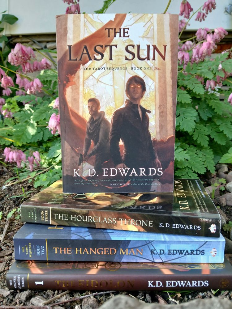 A stack of books in front of blooming bleeding hearts. The book on top, The Last Sun by KD Edwards, is standing on edge, the cover facing the camera. The stack from top to bottom is The Hourglass Throne by K.D Edwards. The Hanged Man by K.D Edwards. The Eidolon by K.D Edwards.