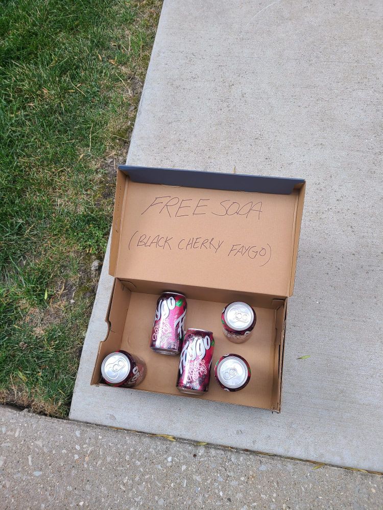 A box of sodas with "FREE SODA (BLACK CHERRY FAYGO)" written in marker across the top sits on a sidewalk