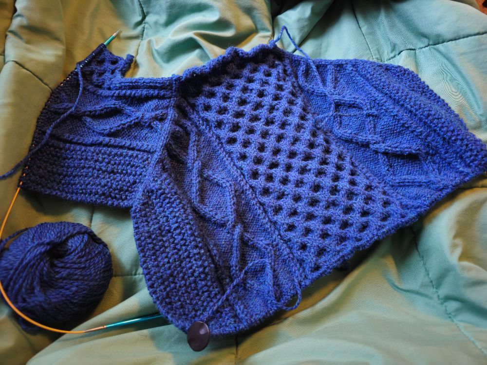 A photo of a cabled sweater being knit. It is the back yoke and left shoulder. It is an ultramarine blue color
