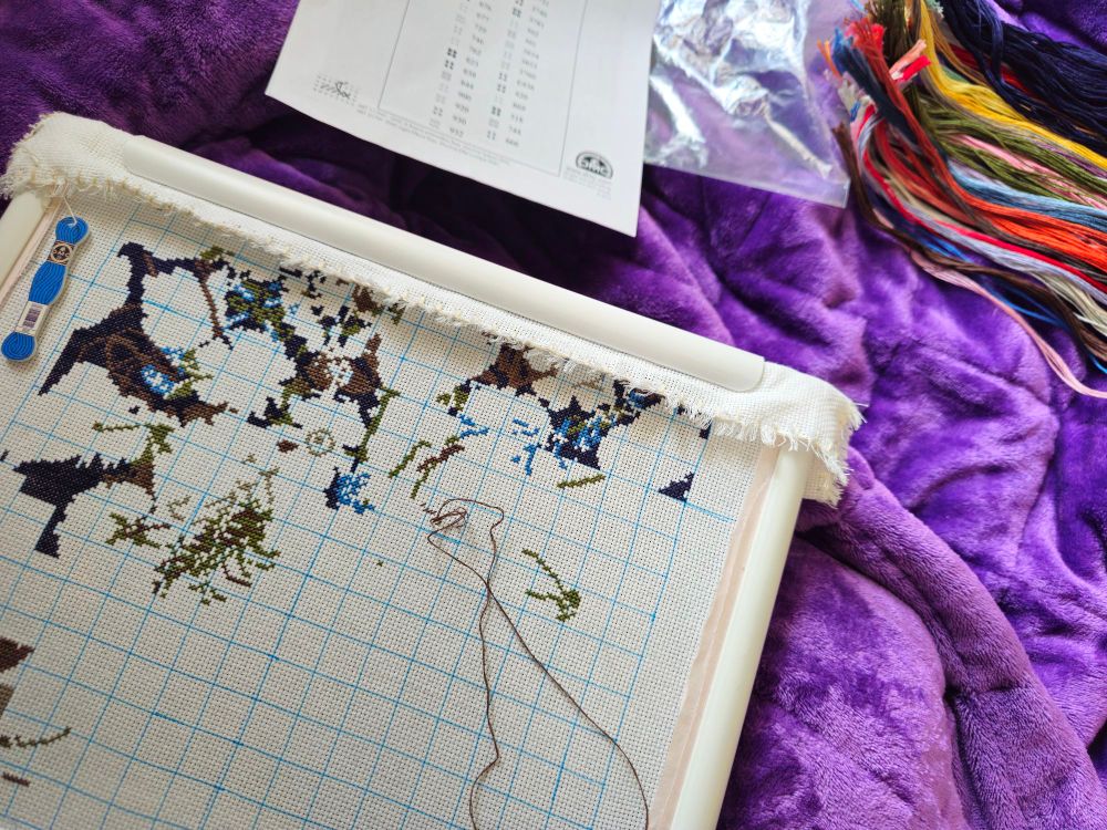 A photo of a cross stitch project in progress. It is sitting on a purple blanket and the chart key and threads are also on the blanket.