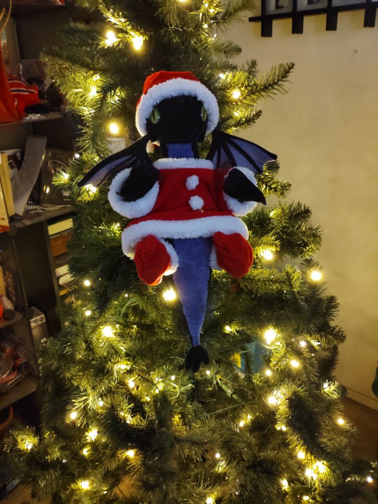A plushie of Malleus Draconia from Twisted Wonderland in his baby dragon form. He's in a Christmas tree and wearing a Santa suit