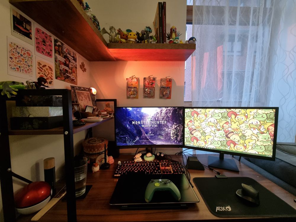 A desk set up with soft orange lighting, and a dual screen monitor set up with monster hunter world loaded on screen one and an illustration of the grass type pokemon starters on the second monitor. A green xbox controller sits atop a drawing display tablet, and pokemon memorabilia sits on a shelf above the desk