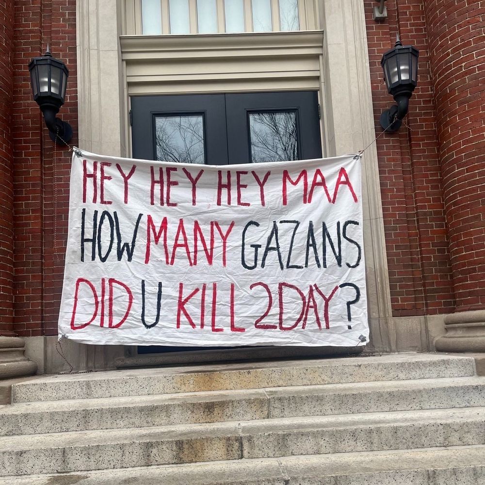 Image of the door of a Harvard building with a big banner--white background, red and black words--that reads Hey Hey Hey MAA / How many Gazans / Did U Kill 2Day? 