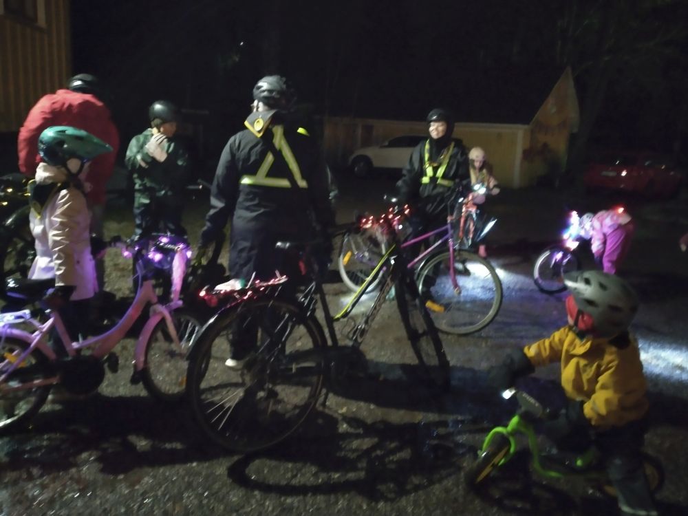 Useita aikuisia ja lapsia koristevalaistujen pyörien kanssa kokoontuneena pihaan, etualalla pieni lapsi potkupyörän kanssa