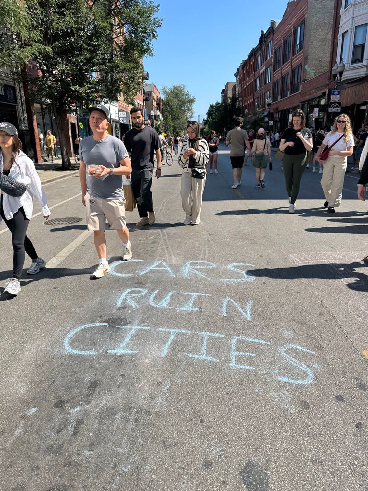 Photo looking southeast down Milwaukee Ave in Chicago on a car free Sunday with chalk wiring on the pavement that says, “CARS RUIN CITIES”