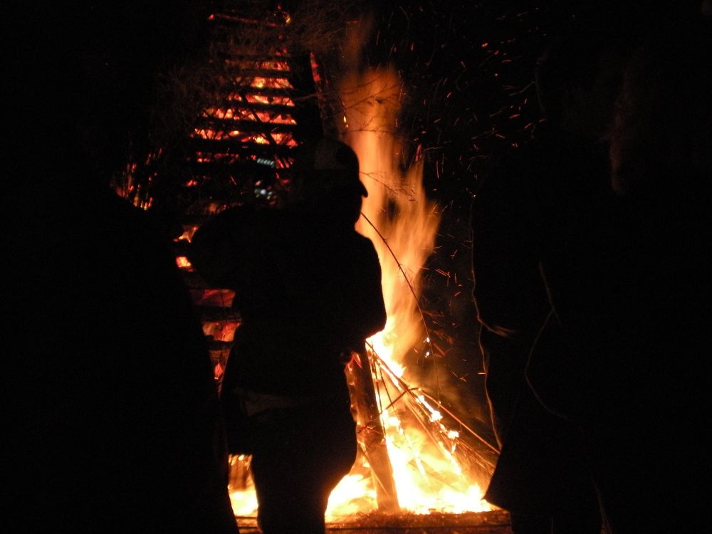 Bonfire ablaze, backlighting person in silhouette with a smartphone.