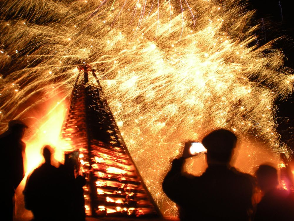 Clash of light with bonfire and fireworks in background. Silhouettes of people in foreground. One man holds up a smartphone, recording the scene.