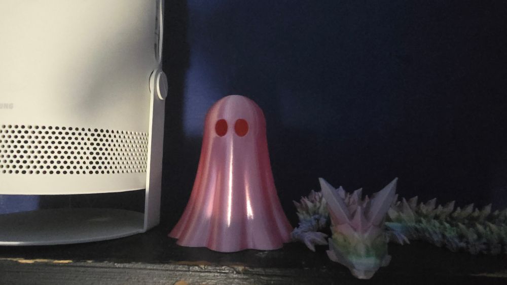 A snaky spiky dragon in rainbow pastel colors and an iridescent pink bedsheet-style ghost, both 3D-printed plastic, sit on a shelf next to a smart projector.