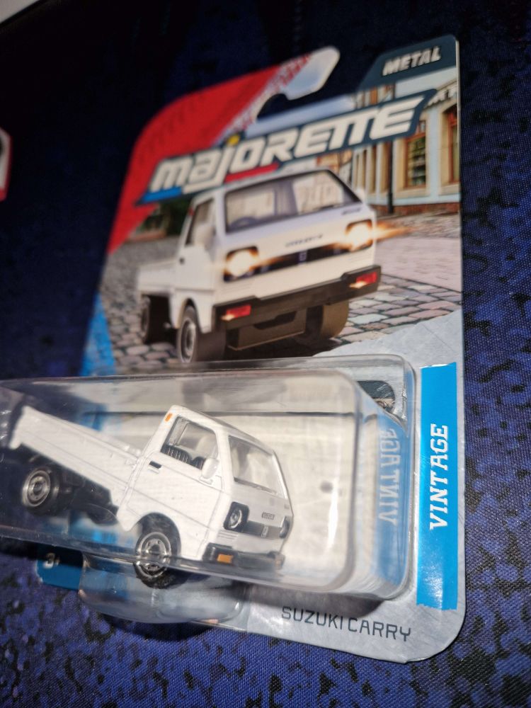 A small metal model car of a Suzuki Carry kei truck
Of the Majorette brand, it's still packaged in a transparent plastic enclosure with an illustrated carboard backing card.