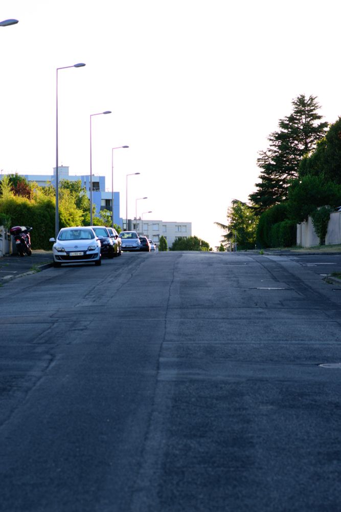 Vertical picture of a street sloping upwards. In the distance, cars are parked on the left side, facing the camera. Trees and hedges are on both side of the street, and residential buildings can be seen further in the background.