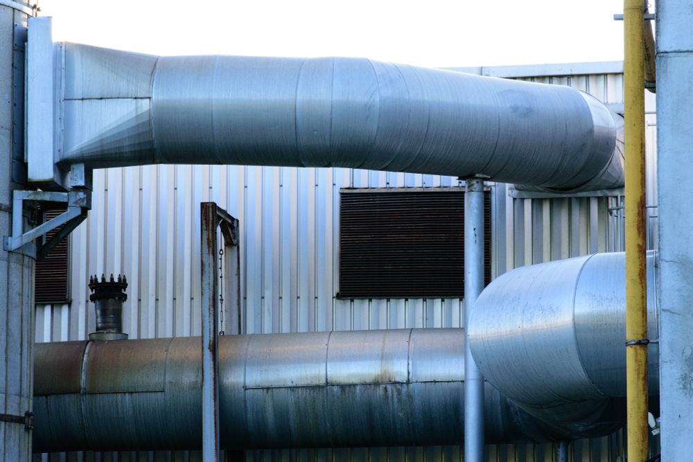 An industrial-looking building. In the background is a metal wall with rusted vents.
The foreground is horizontally crossed by two large metal pipes.

Most of the surfaces are metallic and show some corrosion.
