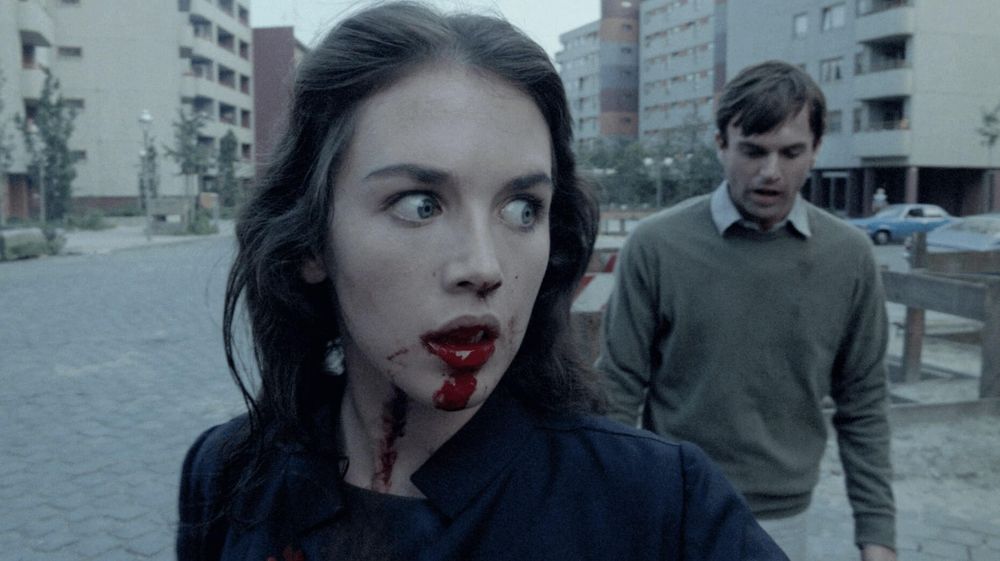 still from Possession 1981 - Isabelle Adjani stares to the right, blood dripping from her open mouth. eyes wide with shock and craze. Sam Neill walks behind her, staring at the ground 