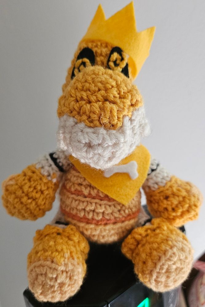 Front view of a crochet dry bones oc plushie wearing a crown and bandana sitting on top of a tower fan.