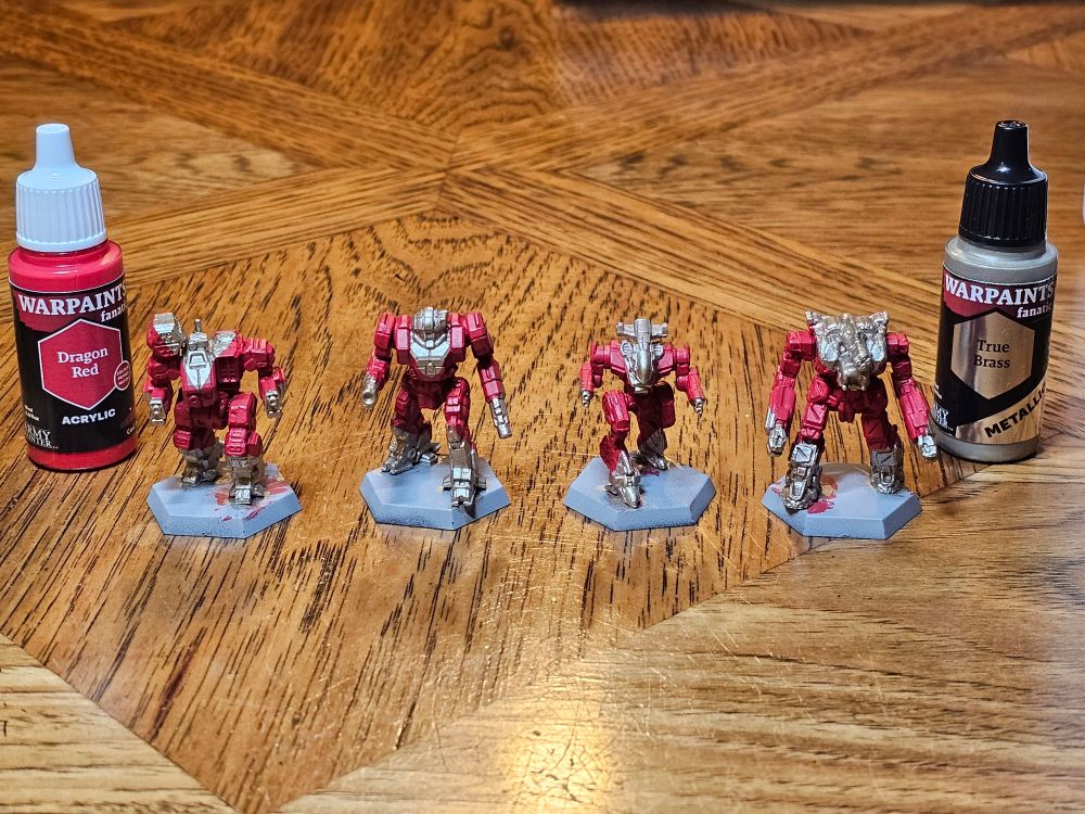From left to right, BattleTech 'Mechs Hellspawn, Enforcer, Gunsmith, and Legionnaire miniatures, with a base coating in 2nd Dragonlords livery of red with brass accents. The paints used to accomplish this, both from the Army Painter's Warpaints Fanatics line, frame the 'Mechs: Dragon Red to thr left and True Brass to the right. 