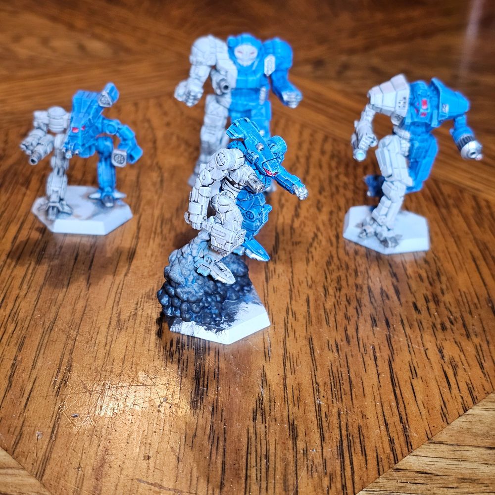 Four BattleTech miniatures painted in 10th Lyran Guards livery. The 'Mechs represented are Hatchetman HCT-6D with jump jets in the foreground and Bushwacker BSW-X1, Atlas AS7-D, and Ymir BWP-2B in the background, from left to right. 

"Lyran Guard equipment is painted blue, with white applied as a broad vertical stripe covering portions of the right torso, arm, and leg."