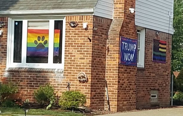 A brick house adorned with a furry Pride flag, a "Trump Won" flag, and a rainbow American flag.