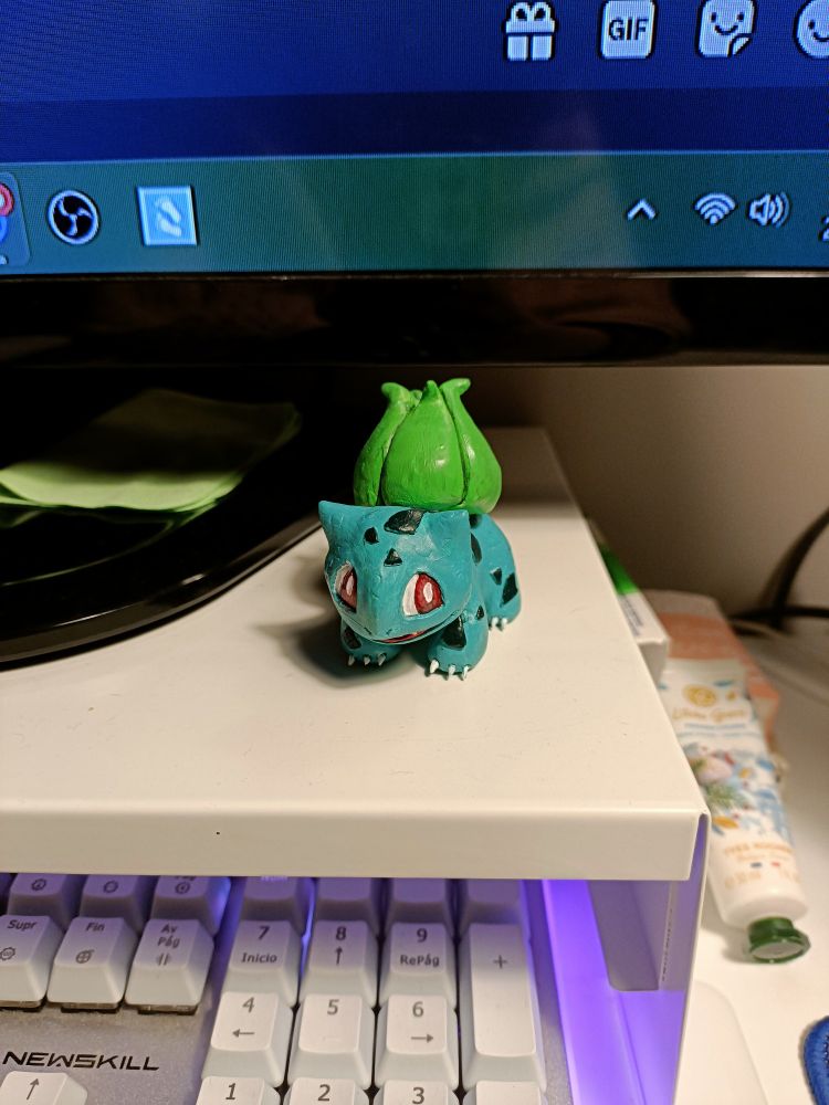Figura artesanal de Bulbasaur, esculpida y pintada a mano. Reposa sobre un elevador de pantalla con un teclado debajo. De frente. 
