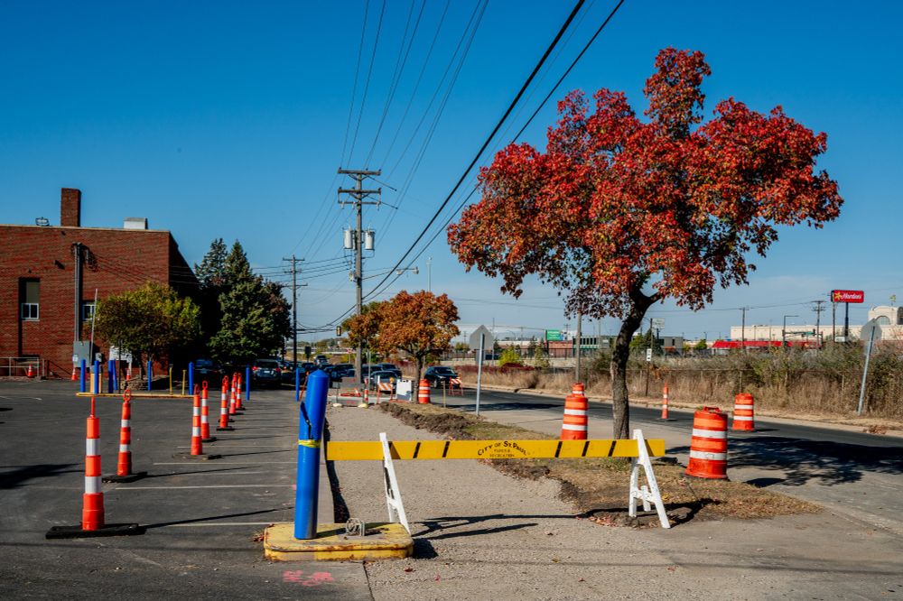 A photograph of a street scene near I-94 in St. Paul, featuring a parking lot lined with bright orange construction barrels and cones. In the center, a large tree with brilliant red autumn leaves stands out. A yellow barricade with 