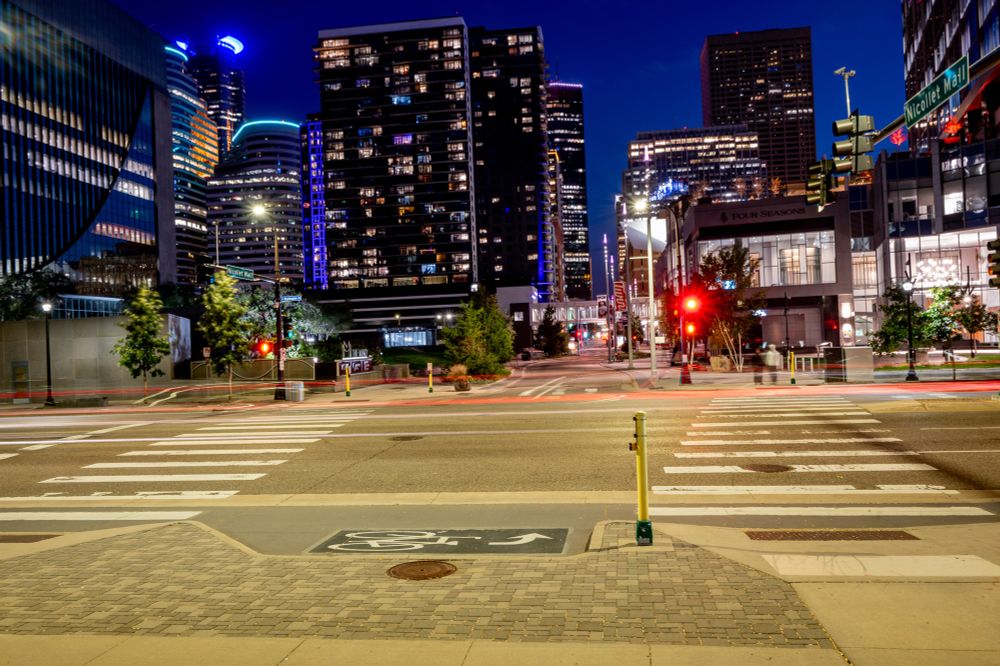 A long-exposure photograph captures a vibrant night scene at the intersection of Nicollet Mall and Washington Ave in downtown Minneapolis. The tall skyscrapers are brightly lit, and the red and white light trails from passing cars are visible on the street. Traffic lights are red, and the crosswalk is clearly marked. The street has a 