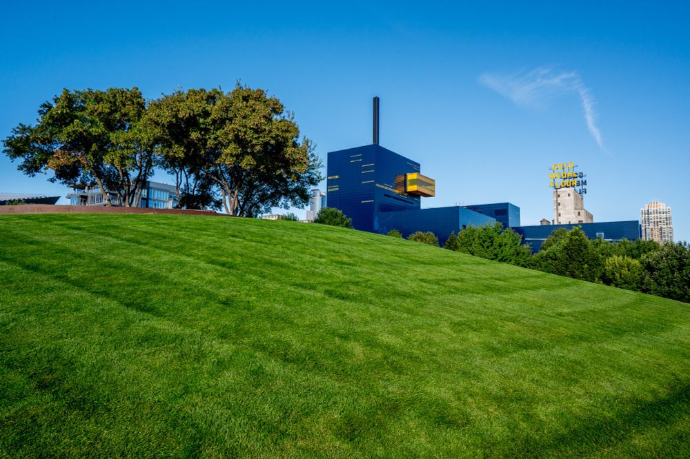 A wide-angle view from Gold Medal Park in downtown Minneapolis shows a large, well-manicured green lawn sloping up towards the top of the frame. In the background, the distinctive dark blue and yellow facade of the Guthrie Theater is visible, along with the nearby 