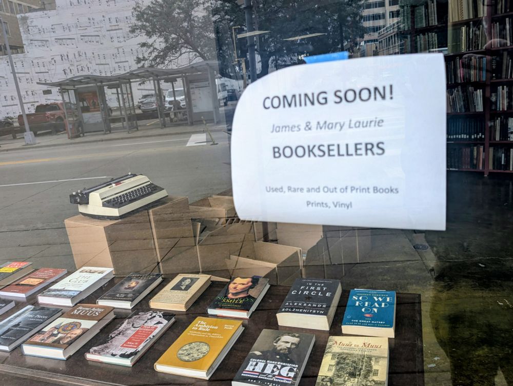 Photo of a coming soon sign for a new used and rare bookstore on a window with books and a typewriter displayed inside the store