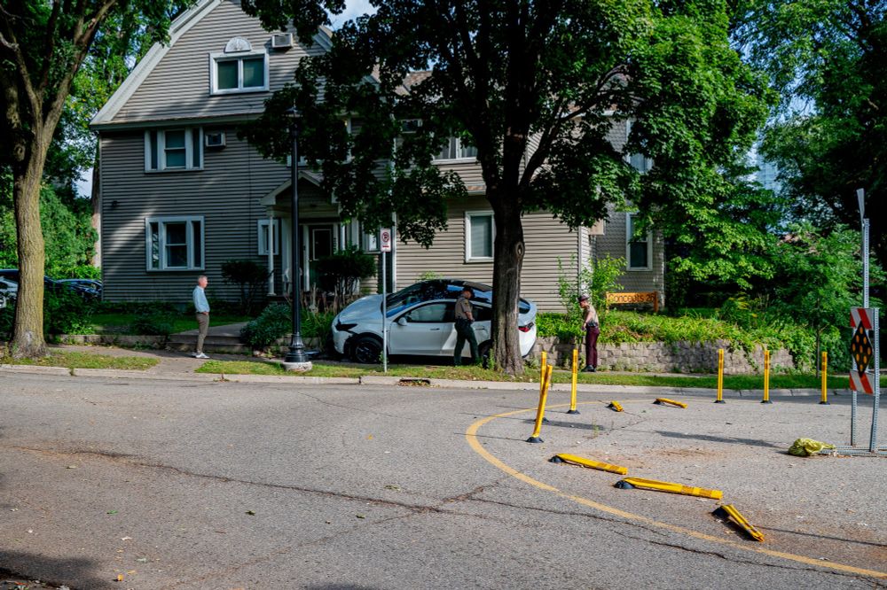 This photograph captures the aftermath of a car accident on a residential street in Minneapolis. A white Tesla has crashed into a small wall and a tree. Two police officers are on the scene, one speaking to a person standing near the vehicle and another speaking to a person in a white shirt. Yellow safety barriers are on the street near the car, indicating the area is marked off.