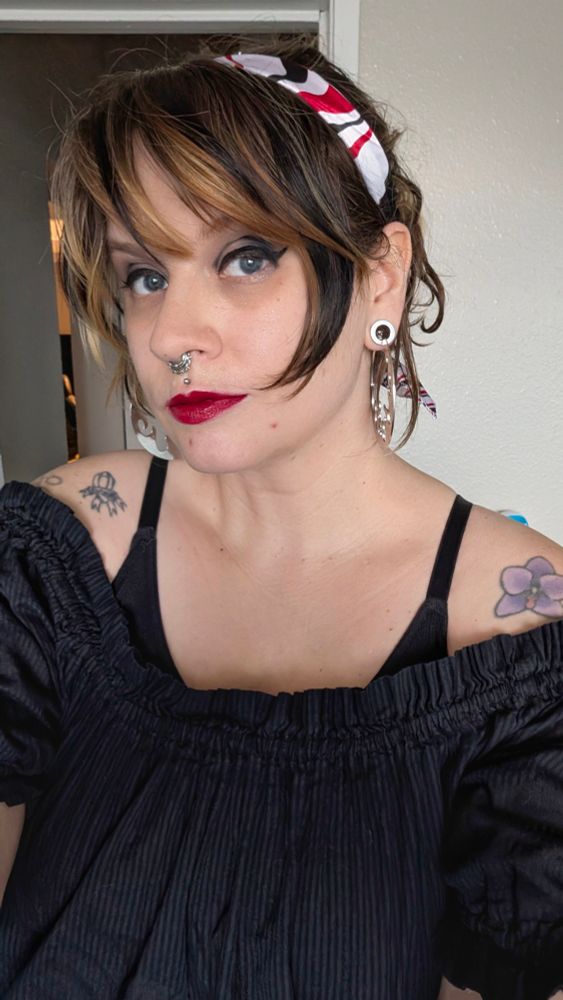 A European American person wearing a black pirate top. Their hair is pulled back. They're wearing a black, red, and white bandana wrap around their head. They have cat eyeliner on and bright red lip stick.