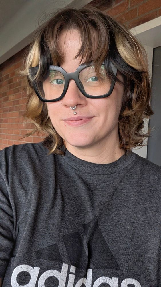 A Caucasian European American wearing thick black framed glasses, wearing a dark heather grey Adidas shirt, Millennial Smirking at the camera. They're standing in front of a brick wall. They have calico colored short hair. They have a Medusa and septum piercing. 