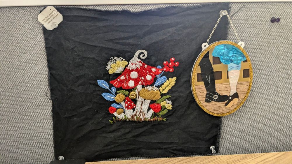 A black piece of fabric with an embroidery of mushroom, flowers, and other plants, thumbtacked onto a cubicle. There's a tea bag in the top left hand corner that says "the finest pleasure is kindness to others", a quote by Jean de La Bruyère. A vinyl plaque of Ed Teach and Stede Bonnet's famous foot touch is hanging off the top right hand thumbtack. 