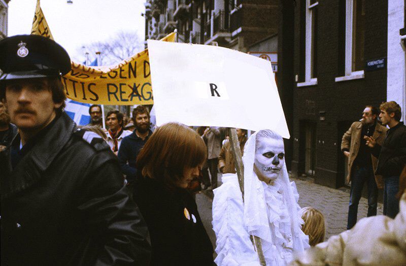 На фотографии изображено костюмированное шествие по центру какого-то европейского города. Активисты несут плакаты и транспаранты на германском языке. Дама, изображающая скелет в белом одеянии или белую смерть, держит плакат на тему «радиоактивной смерти». Её белый плакат заретуширован, на нём осталась только одинокая буква R. По цветопередаче кадра, а также по одежде и причёскам протестующих можно предположить, что фотография сделана в конце XX века. // The photograph depicts a costumed procession through the centre of a European city. Activists carry placards and banners in German. A lady, portraying a white-robed skeleton or white death, holds a banner on the topic of “radioactive death.” Her white banner has been retouched, revealing only a single letter R. The colour rendition, as well as the clothing and hairstyles of the protesters, suggest that the photograph was taken in the late 20th century. // © Roel Burgler