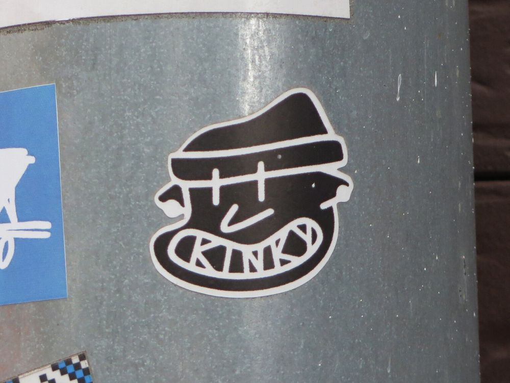 Чёрно-белый стикер в виде головы маскулинного персонажа в вязаной шапке. Нос-галочка похож на логотип «Найка», в обоих ушах тоннели или кольца, большой рот широко раскрыт и содержит внутри себя надпись Kinky. // A black and white sticker depicting the head of a masculine character wearing a knitted hat. The nose is shaped like a Nike logotype, both ears have tunnels or rings, and the large mouth is wide open and contains the name “Kinky” inside. // 2025 © Kinky